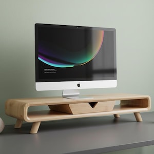 May include: A light-colored wooden monitor stand with a built-in drawer supports a silver iMac computer. A small succulent in a concrete pot and a ceramic mug sit on the desk. The computer screen displays a colorful abstract design.
