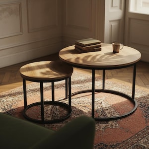 May include: Two round wooden side tables with black metal legs. The tables are different sizes, with the larger one holding books and a mug. They sit on a patterned rug in a room with natural light.