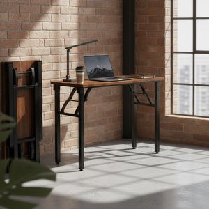 May include: A rectangular desk with a dark wood-grain surface and a black metal frame. The desk features foldable legs and is positioned next to a folded version of the same desk. A laptop, a desk lamp, and a coffee cup are placed on the desk.
