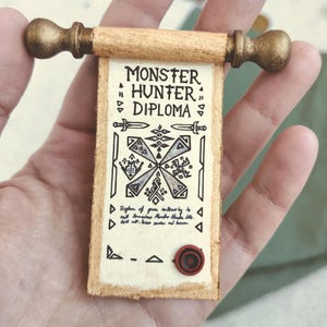 Op de afbeelding: Een miniatuurrol met de tekst "MONSTER HUNTER DIPLOMA" in zwarte inkt. De rol is gemaakt van bruin papier met een decoratief ontwerp en een rode zegel. De rol wordt vastgehouden door een goudkleurige houten handgreep.