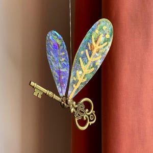 May include: A hanging ornament featuring an antique brass-coloured skeleton key with two iridescent, translucent wings attached. The wings display a shimmering, multi-coloured texture with a gold-painted leaf vein pattern on the larger wing.