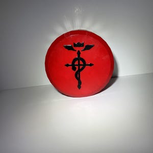 May include: A circular red disc featuring a raised black emblem in the centre. The emblem consists of a cross with a serpent coiled around it, topped by a small crown and two wings. The surface of the red disc has a textured, layered appearance.