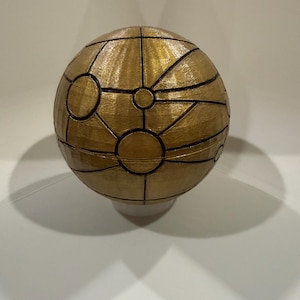 May include: A spherical, gold-colored decorative object with a geometric design. The sphere is divided into sections by black lines, with circular and linear patterns. The object is resting on a white base, casting a shadow on the white surface.