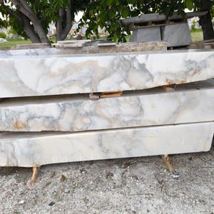 Può includere: Tre lastre di marmo impilate con base bianca e venature grigie e beige. Le lastre rettangolari sono all'aperto, appoggiate su supporti in legno. Il marmo ha una finitura lucida, che mette in risalto i motivi naturali.