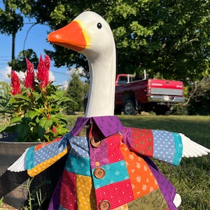 May include: A white goose statue with an orange beak wearing a colorful patchwork coat. The coat features various fabric patterns in shades of red, orange, yellow, blue, and purple, secured with three brown buttons down the front center.