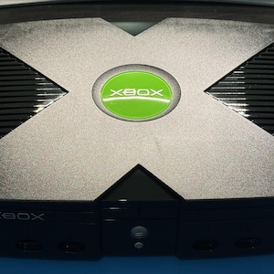 May include: A black original Xbox video game console featuring a large X-shaped top panel with a circular green and silver Xbox logo in the centre. The front face includes the Xbox text logo, controller ports, and a power button in a black housing.
