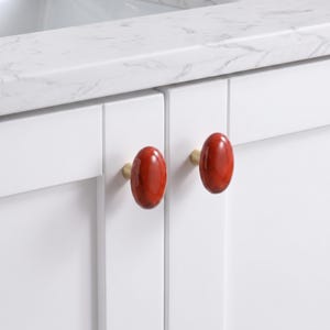 May include: Two oval-shaped cabinet knobs with a glossy, deep red marbled finish and gold-toned metal bases. These smooth, rounded knobs are mounted onto white cabinet doors, providing a distinct contrast against the clean, neutral surface.