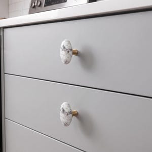 May include: Two oval-shaped cabinet knobs featuring a white and grey marble pattern with dark veining. Each knob is attached to a light grey drawer front by a small, cylindrical brass-toned metal base that connects the marble to the drawer surface.