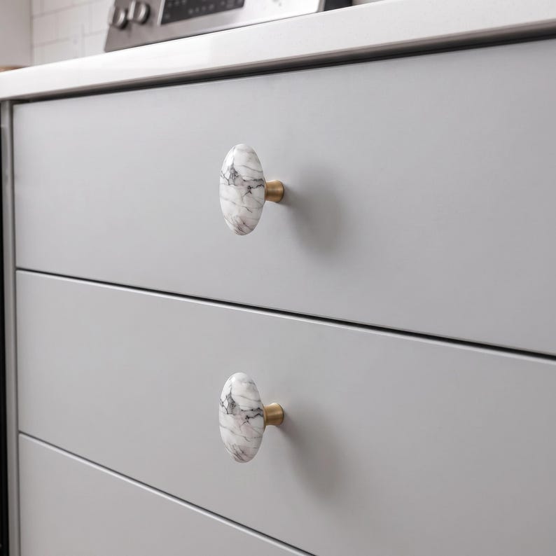 May include: Two oval-shaped cabinet knobs featuring a white and grey marble pattern with dark veining. Each knob is attached to a light grey drawer front by a small, cylindrical brass-toned metal base that connects the marble to the drawer surface.