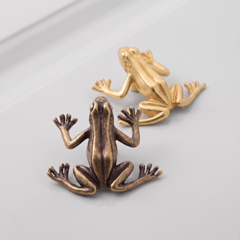 May include: Two metal frog-shaped cabinet knobs. One knob features a bright, polished gold finish, while the other displays a darker, antique bronze finish. Both knobs are depicted in a flat, splayed position with visible limbs and webbed feet.