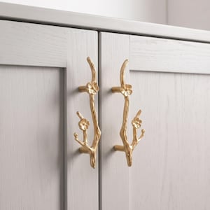 May include: Two vertical gold-toned cabinet pulls shaped like slender tree branches with small, five-petaled flowers attached. Each handle is mounted onto a light gray wooden cabinet door with a visible grain, featuring a smooth, metallic finish.