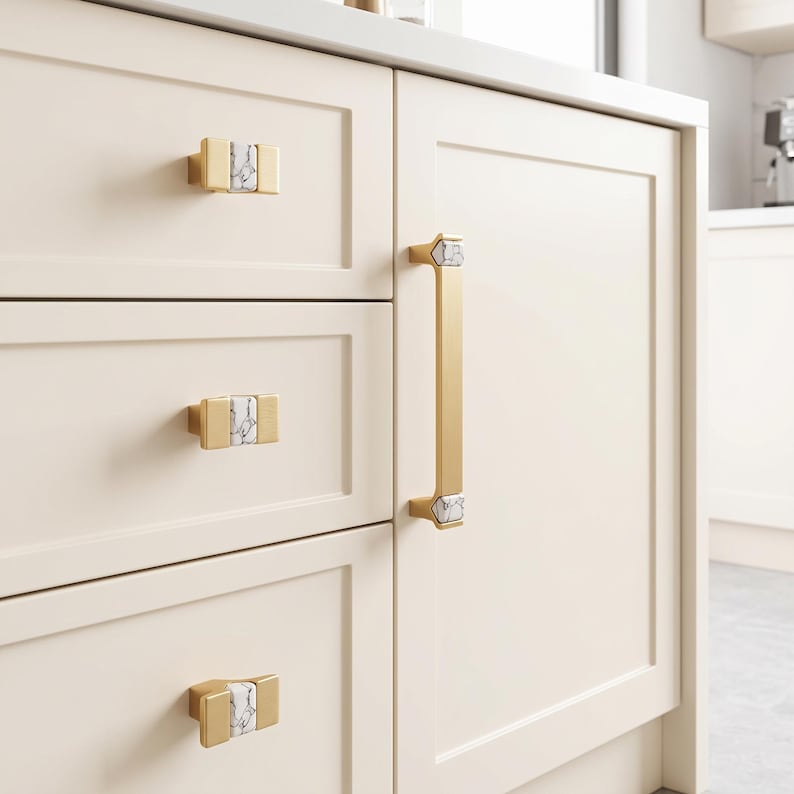 Peut inclure: Un ensemble de poign&eacute;es de placard dor&eacute;es comprenant trois boutons rectangulaires et une longue poign&eacute;e verticale. Chaque pi&egrave;ce est orn&eacute;e d'une incrustation centrale en marbre blanc et gris, fix&eacute;e sur des meubles de cuisine cr&egrave;me &agrave; panneaux.