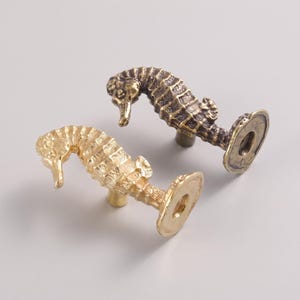 Puede incluir: Dos pomos para armarios con forma de caballito de mar hechos de metal. Un pomo presenta un acabado dorado brillante y pulido, mientras que el otro tiene un acabado de bronce antiguo más oscuro. Cada pomo tiene un cuerpo texturizado y una base.