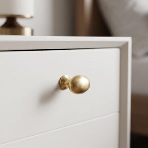 May include: A brushed gold, oval-shaped cabinet knob attached to a smooth, off-white drawer front. The hardware features a rounded, metallic surface with a soft satin finish, centred on the flat panel of the furniture piece.