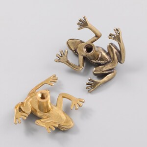 May include: Two brass-toned metal drawer knobs shaped like frogs. Each knob features a detailed frog figure with outstretched limbs and a central threaded cylindrical post on the underside for mounting, showing a metallic finish and texture.