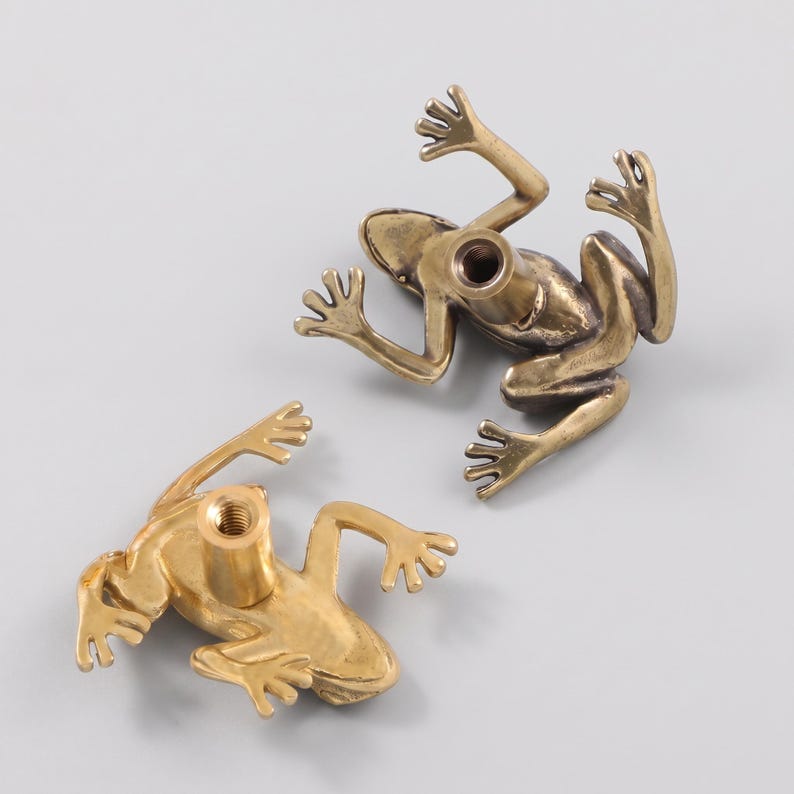 May include: Two brass-toned metal drawer knobs shaped like frogs. Each knob features a detailed frog figure with outstretched limbs and a central threaded cylindrical post on the underside for mounting, showing a metallic finish and texture.