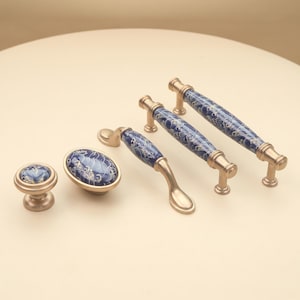 May include: A collection of four cabinet hardware pieces featuring a brushed gold metal finish and a central blue ceramic inlay decorated with a white swirling wave pattern. The set includes one round knob, one oval knob, and two varying lengths of drawer pulls.