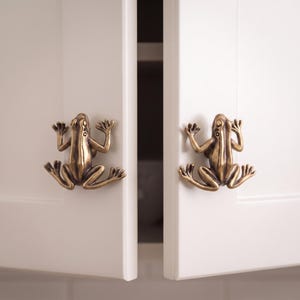 May include: Two brass-colored cabinet knobs shaped like frogs with detailed limbs and eyes. Each knob is mounted on a white cabinet door, featuring a metallic finish with dark highlights that accentuate the sculpted form of the amphibian design.