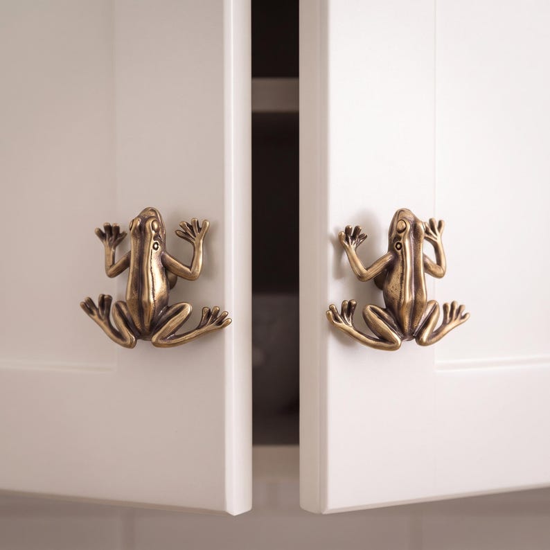 May include: Two brass-colored cabinet knobs shaped like frogs with detailed limbs and eyes. Each knob is mounted on a white cabinet door, featuring a metallic finish with dark highlights that accentuate the sculpted form of the amphibian design.