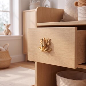 May include: A light-colored wooden drawer front featuring a metallic gold-toned frog-shaped handle. The drawer is partially open, revealing the smooth grain of the wood and the detailed, sculpted form of the frog hardware attached to the center.