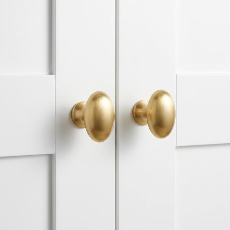 May include: Two oval-shaped cabinet knobs with a smooth, brushed gold finish. Each knob is mounted onto a white cabinet door panel with a vertical seam running between them. The hardware features a rounded, symmetrical design with a metallic sheen.