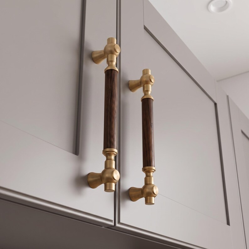 Pu&ograve; includere: Due maniglie verticali per armadietti con una sezione centrale cilindrica in legno marrone scuro, accentuata da ferramenta in ottone spazzolato nella parte superiore e inferiore. La ferramenta include basi di montaggio circolari su armadi grigio chiaro.