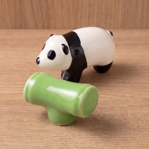 May include: A ceramic panda figurine with a glossy white and black finish, positioned behind a separate, smaller, cylindrical green ceramic piece that resembles a segment of bamboo. The panda has black ears, eye patches, and legs.