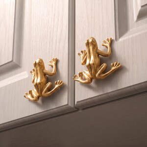 May include: Two gold-colored cabinet knobs shaped like frogs, each featuring a smooth, metallic finish and detailed limbs. The knobs are mounted onto light gray cabinet doors with a visible wood grain texture, serving as functional hardware.