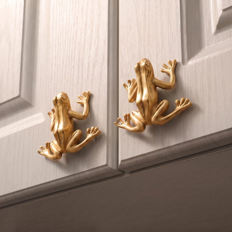May include: Two gold-colored cabinet knobs shaped like frogs, each featuring a smooth, metallic finish and detailed limbs. The knobs are mounted onto light gray cabinet doors with a visible wood grain texture, serving as functional hardware.