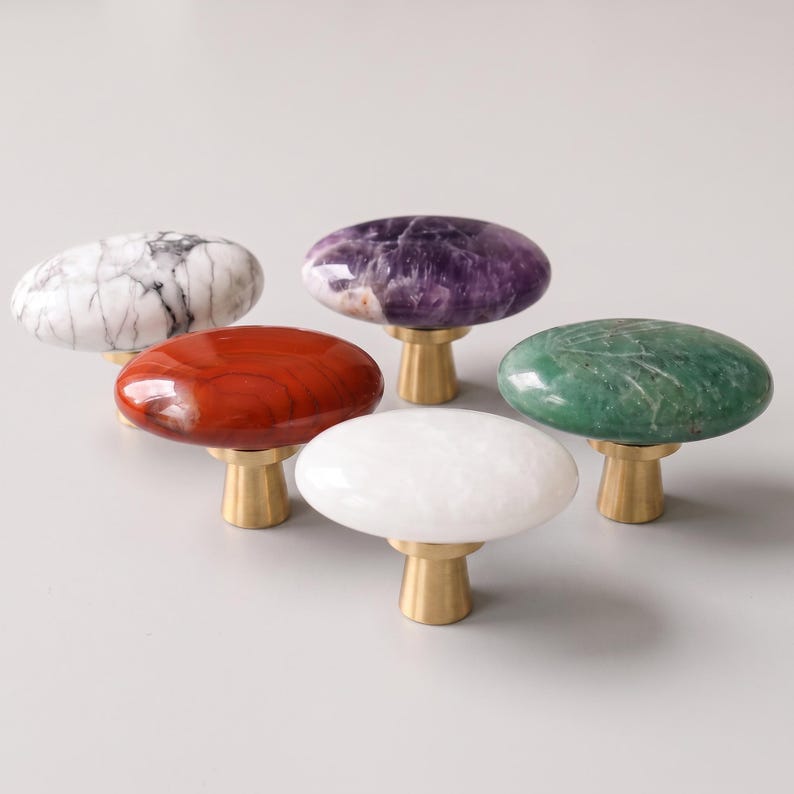 May include: Five cabinet knobs with smooth, oval-shaped stone tops in white marble, purple amethyst, red jasper, solid white, and green aventurine. Each knob features a polished brass-toned metal base with a tapered, cylindrical design.