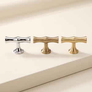 May include: Three T-shaped cabinet knobs arranged in a row. The first knob on the left has a polished chrome finish. The middle knob features a brushed antique brass finish, and the third knob on the right has a smooth, satin gold metal finish.