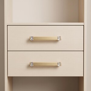Peut inclure: Deux tiroirs en bois de couleur claire dot&eacute;s de poign&eacute;es horizontales en m&eacute;tal dor&eacute; bross&eacute;. Chaque poign&eacute;e est agr&eacute;ment&eacute;e d'&eacute;l&eacute;ments en verre transparent, rectangulaires et facett&eacute;s aux deux extr&eacute;mit&eacute;s, ajoutant une finition d&eacute;corative.