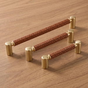 May include: Three cabinet pulls of varying lengths, each featuring a central handle wrapped in brown braided material and finished with cylindrical brushed brass end caps. The hardware is arranged diagonally to show the different sizes.