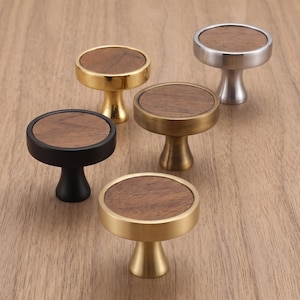 Può includere: Cinque pomelli circolari per armadietti con un inserto piatto in legno sulla parte superiore. I pomelli presentano diverse finiture metalliche, tra cui oro lucido, nero opaco, ottone spazzolato e argento spazzolato, con una base svasata.