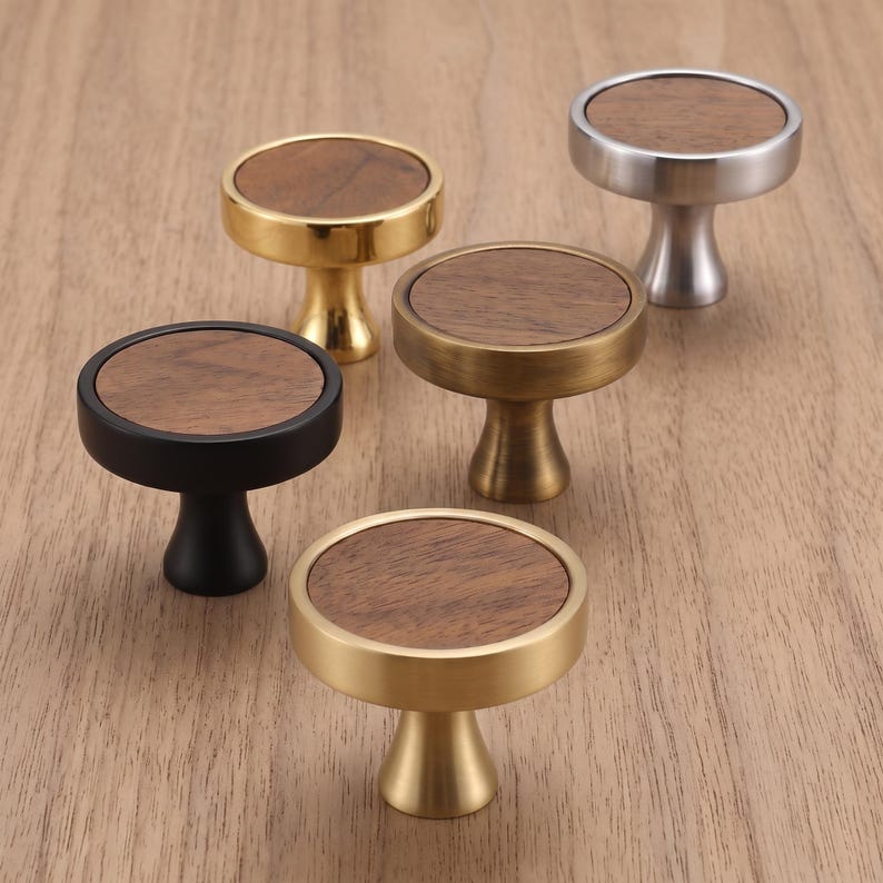 May include: Five circular cabinet knobs with a flat wooden top inlay. The knobs feature different metal finishes including polished gold, matte black, brushed brass, and brushed silver, each with a flared base and a circular wooden centre.