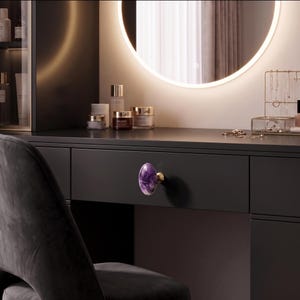 May include: A round, polished purple amethyst stone drawer knob with a gold-toned metal base, mounted on a matte black drawer front. The stone features natural white and deep violet marbling patterns throughout its smooth, rounded surface.