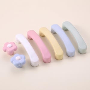 May include: A collection of furniture hardware including two flower-shaped knobs in pink and light blue, and five arched drawer pulls in white, pink, yellow, light purple, and light green. Each piece features a smooth, matte, solid-colored finish.