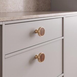 May include: Two circular cabinet knobs featuring a brushed gold metal rim and a dark brown wood grain inlay. The knobs are mounted on a light grey drawer front, each displaying a smooth, flat face with a distinct vertical wood grain pattern.