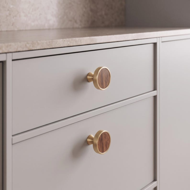 May include: Two circular cabinet knobs featuring a brushed gold metal rim and a dark brown wood grain inlay. The knobs are mounted on a light grey drawer front, each displaying a smooth, flat face with a distinct vertical wood grain pattern.