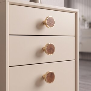 May include: A light beige three-drawer chest featuring circular cabinet knobs. Each knob has a metallic gold-toned rim surrounding a circular wooden inlay with a visible grain pattern. The chest has a smooth, matte finish and clean, straight edges.