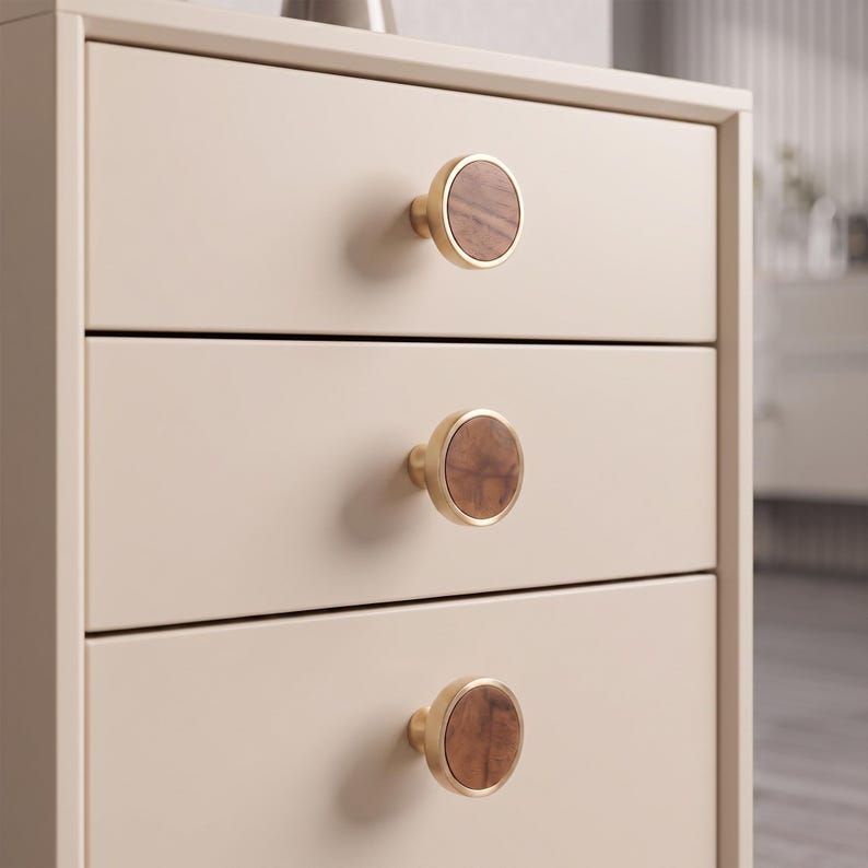 May include: A light beige three-drawer chest featuring circular cabinet knobs. Each knob has a metallic gold-toned rim surrounding a circular wooden inlay with a visible grain pattern. The chest has a smooth, matte finish and clean, straight edges.