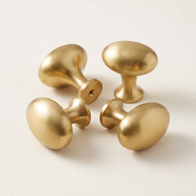 May include: Four oval-shaped cabinet knobs with a brushed gold finish. Each knob features a smooth, rounded top surface and a tapered base that connects to a central mounting point with a visible threaded hole for installation hardware.