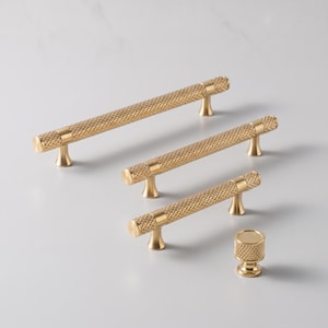 May include: A collection of gold-toned cabinet hardware including three cylindrical bar pulls of varying lengths and one circular knob. Each piece features a textured, diamond-patterned knurled surface with smooth, polished end caps and mounting posts.