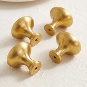 May include: Four mushroom-shaped cabinet knobs crafted from brushed brass metal. Each knob features a smooth, rounded top and a tapered base with a central threaded hole for mounting, showcasing a consistent matte gold finish across all pieces.