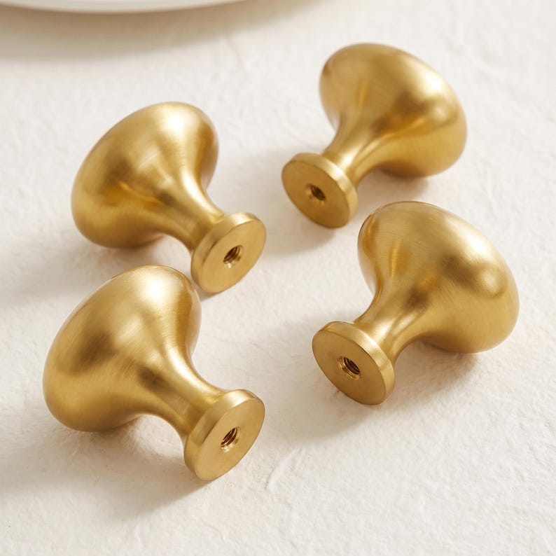 May include: Four mushroom-shaped cabinet knobs crafted from brushed brass metal. Each knob features a smooth, rounded top and a tapered base with a central threaded hole for mounting, showcasing a consistent matte gold finish across all pieces.