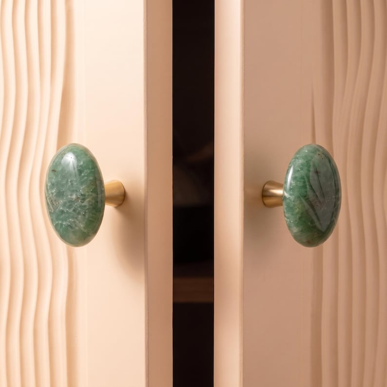 May include: Two oval-shaped green stone cabinet knobs with a polished finish, each mounted on a small gold-toned metal base. The knobs are attached to two light beige cabinet doors featuring a vertical wavy, textured surface pattern.