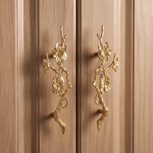 May include: Two vertical cabinet handles made of polished gold-toned metal. Each handle features a sculptural design of a slender branch with two blooming flowers attached. The handles are mounted onto a light brown wooden cabinet door surface.