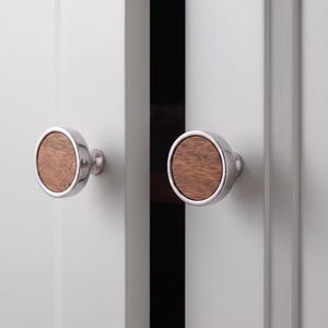 May include: Two circular cabinet knobs featuring a polished chrome metal outer ring and a flat, circular brown wood grain inlay centre. The knobs are mounted on two adjacent white cabinet doors with a vertical gap between them.