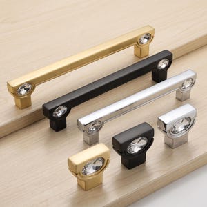 Könnte beinhalten: Eine Kollektion von Möbelbeschlägen, bestehend aus drei langen, rechteckigen Schubladengriffen und drei Knöpfen mit einem Loch. Die Beschläge sind in Gold, Mattschwarz und poliertem Silber erhältlich, jeweils mit einem runden Kristallakzent.