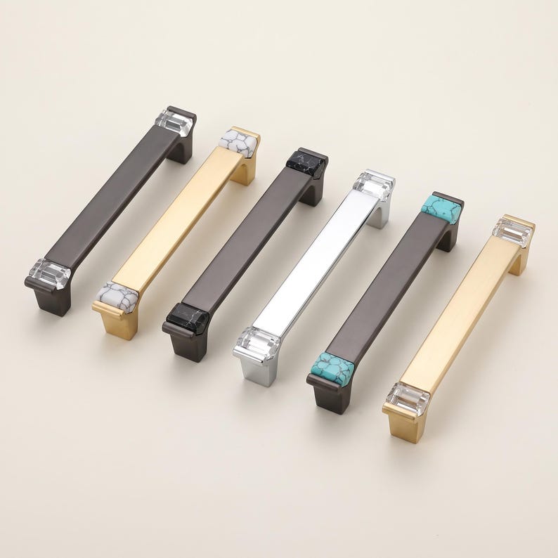 Peut inclure: Six poign&eacute;es de placard rectangulaires dispos&eacute;es en rang&eacute;e. Les poign&eacute;es pr&eacute;sentent des finitions m&eacute;talliques noir mat, or bross&eacute; et argent poli. Chaque poign&eacute;e est orn&eacute;e d'embouts d&eacute;coratifs en cristal transparent, marbre blanc ou turquoise.